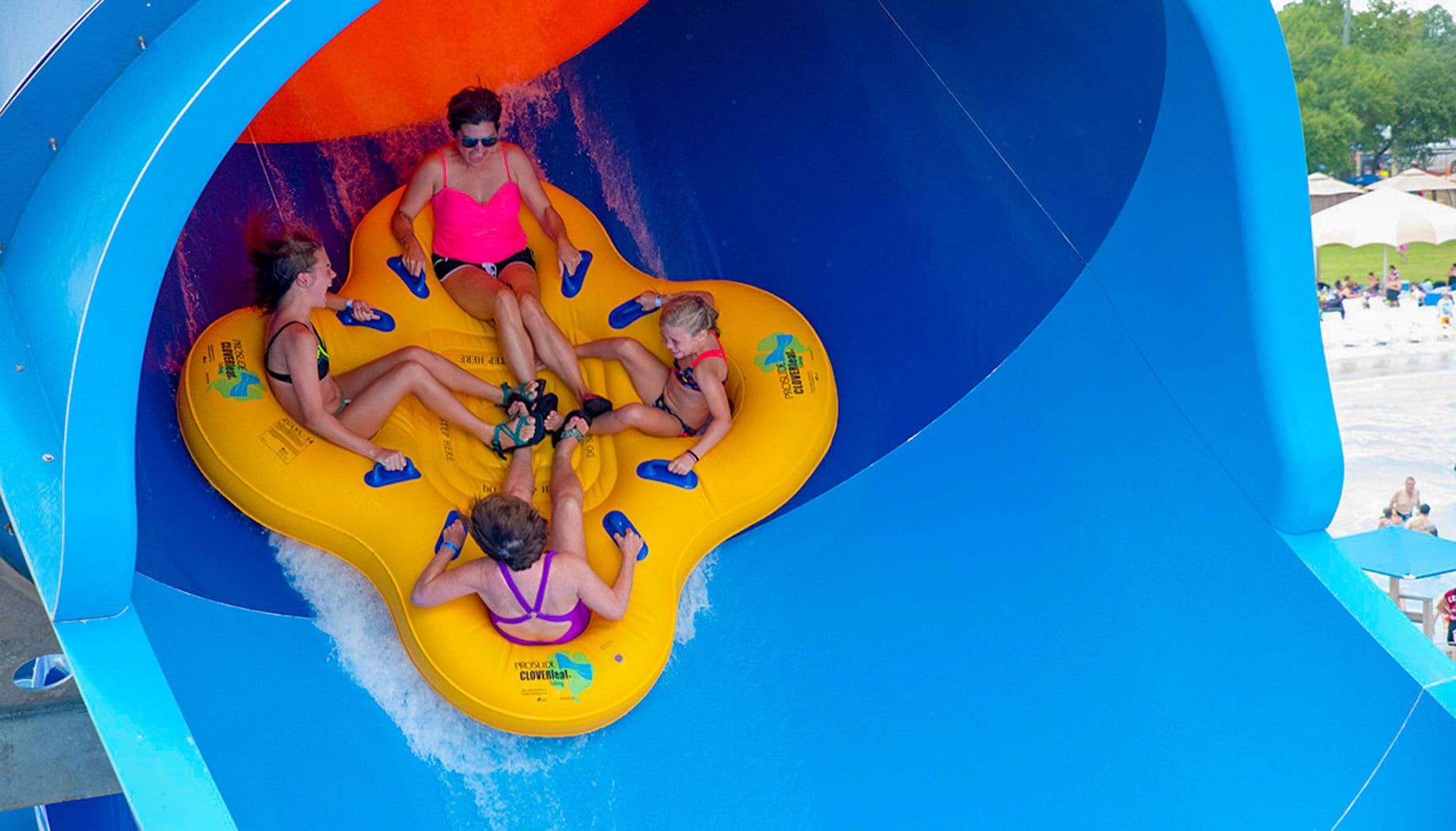 Four people are riding a bright yellow inflatable raft down a blue water slide at a water park.